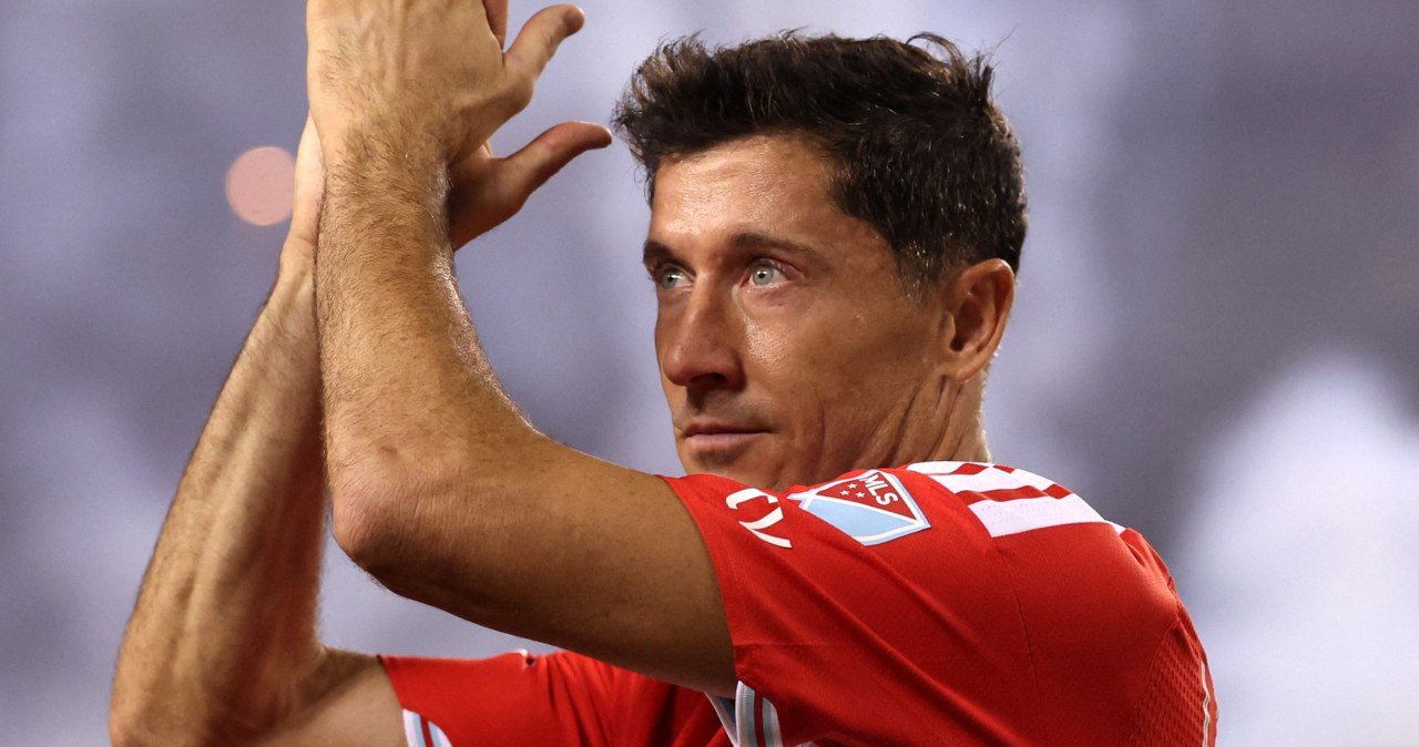 Robert Lewandowski w stroju Chicago Fire z uniesionymi dłońmi, patrzy przed siebie na boisku