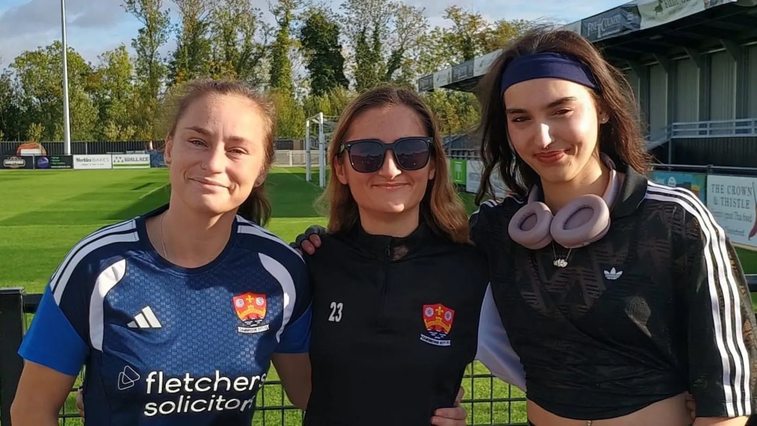 Tom Jackson/BBC Left to right are Shelbee Clarke, Izzy Papandronicou and Marni Voak. They are standing close together in front of a football pitch and are wearing football shirts.
