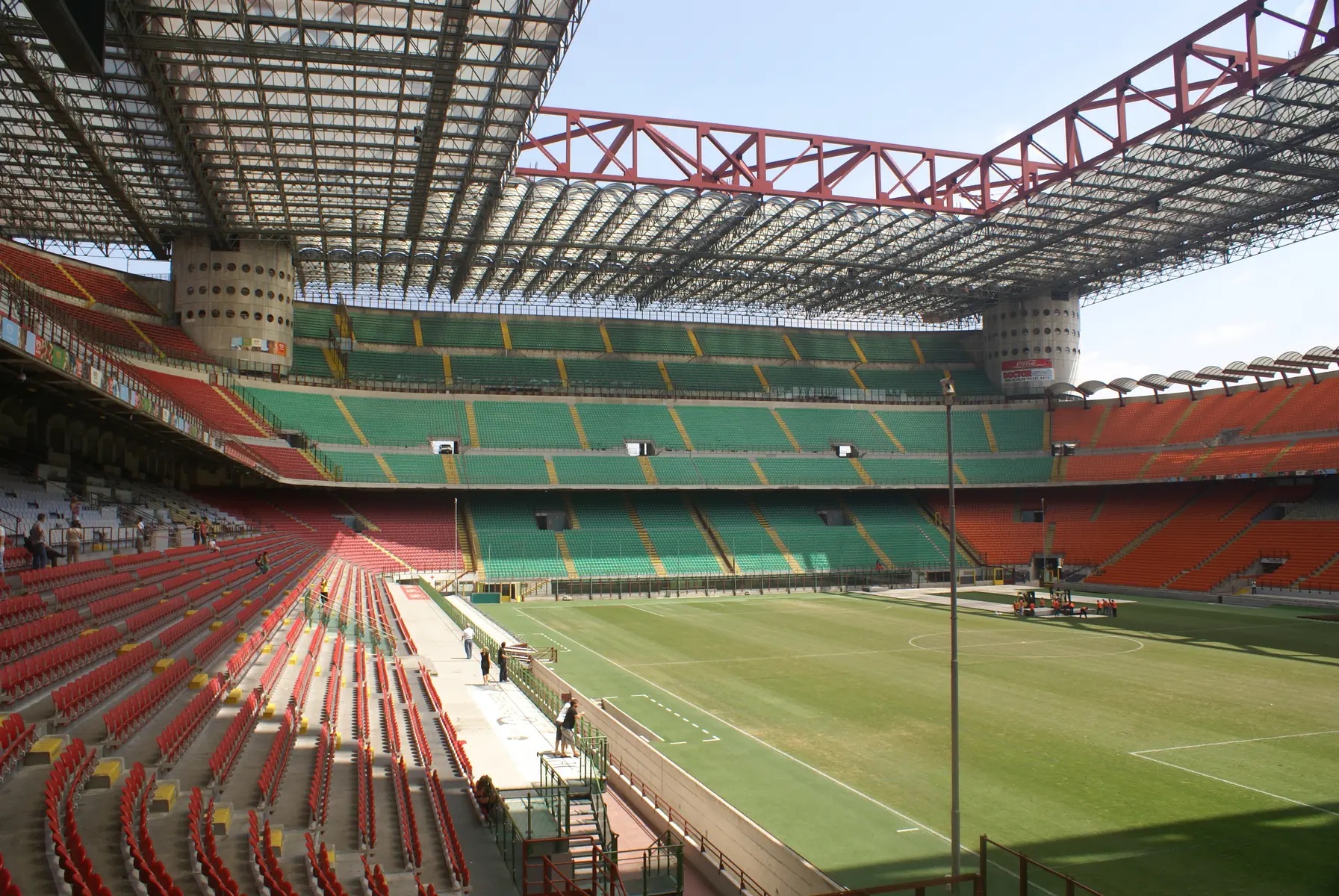 A wide panorama of the empty San Siro stadium in Milan, showing the green and red seating and the overhead roof structure.