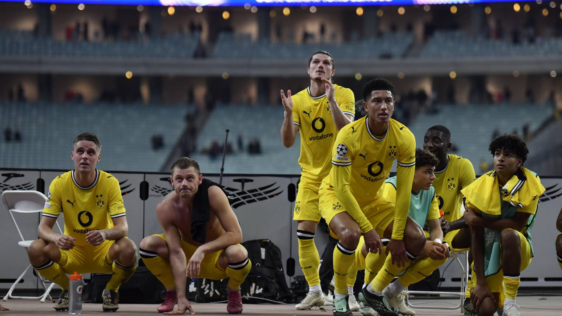 Jogadores de futebol do Borussia Dortmund, alguns agachados e outros em pé, observam o campo. Vestem uniformes amarelos e pretos, com um jogador sem camisa e uma toalha no ombro. Estão à beira do campo, com arquibancadas ao fundo.