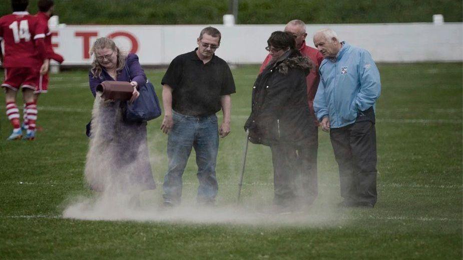 People stood on a football pitch drop ashes from an urn onto the pitch. There are five people in a group and one woman looks to be upset
