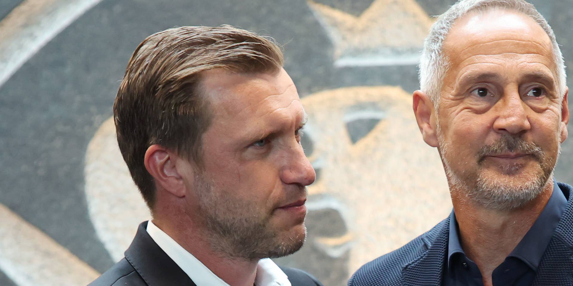 Zwei Männer in Anzügen stehen nebeneinander vor einem Wandmotiv mit Adler.