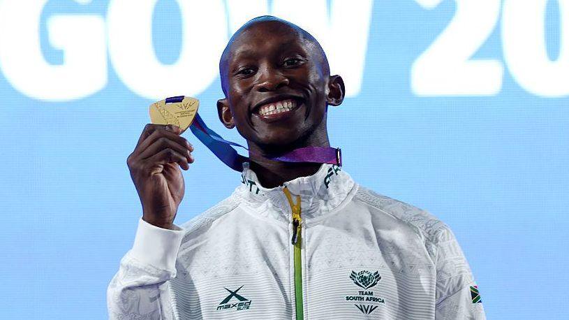 Sinesipho Dambile, visible from chest up wearing a white South African tracksuit top with green zip and trim, poses on a podium with his 2026 Commonwealth Games 200m gold medal.