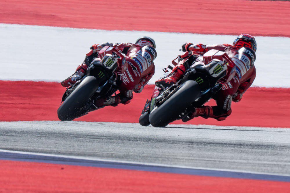 Two riders in red Ducati leathers lean into a corner on MotoGP bikes, with Monster Energy logos visible on the fairings.