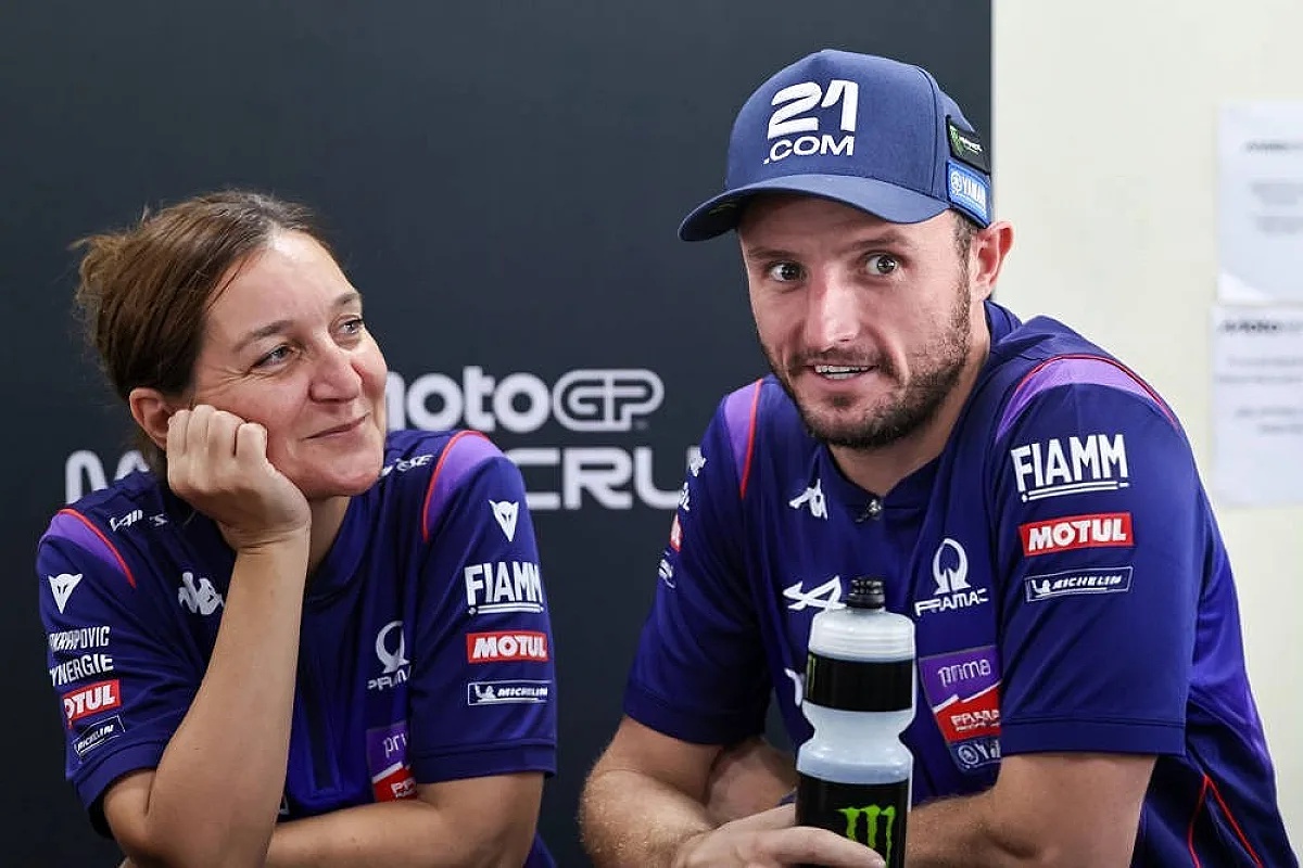 Two people in purple Pramac-style MotoGP team shirts at a press event; a man in a 21.com cap leans on a table beside a Monster bottle.