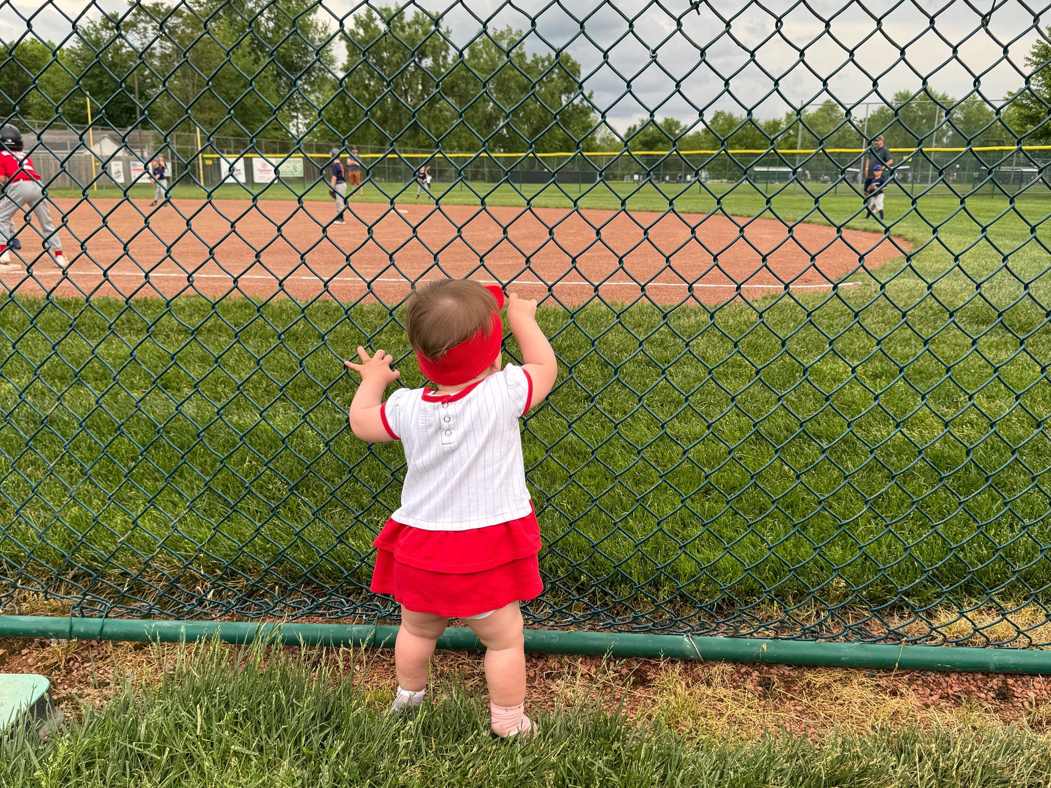 A spectator in red and white stands at a chain-link fence watching a baseball game on a grassy field.