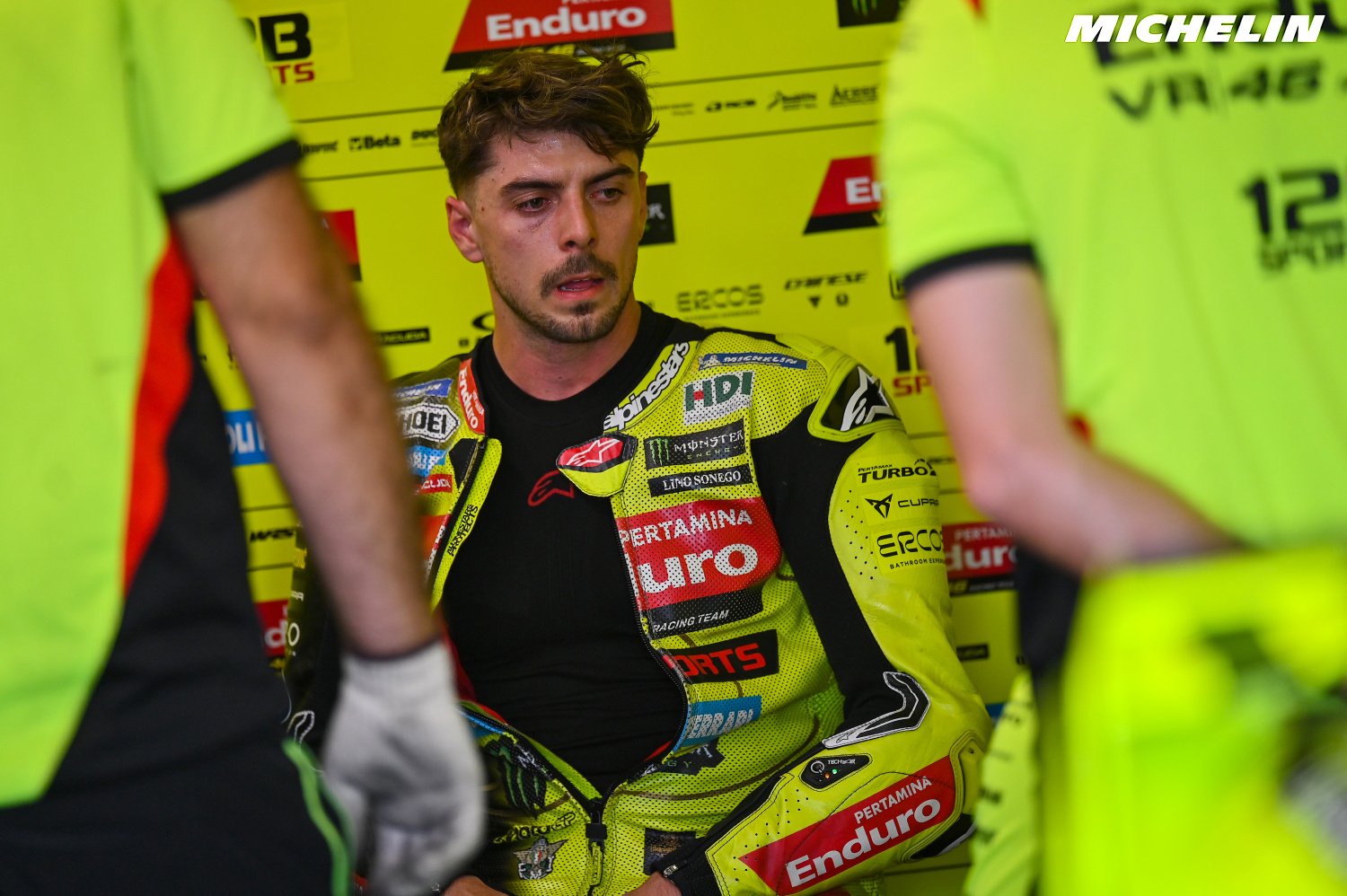 Rider in yellow Pertamina Enduro leathers sits in a MotoGP paddock garage, with teammates in yellow suits beside him