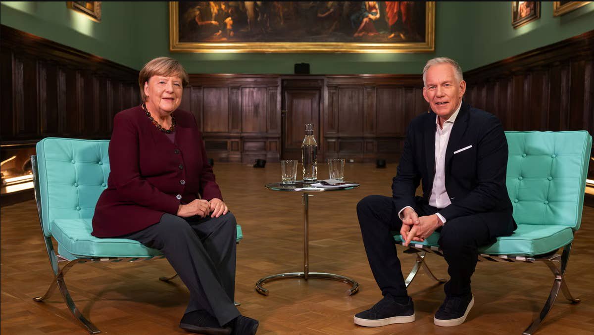 Angela Merkel und Johannes B. Kerner sitzen in einem eleganten Raum auf türkisfarbenen Sesseln.