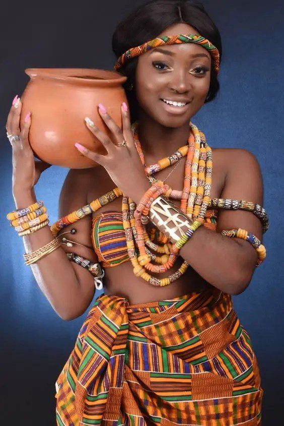 A lady dressed in kente fabric with beads, carrying a clay pot