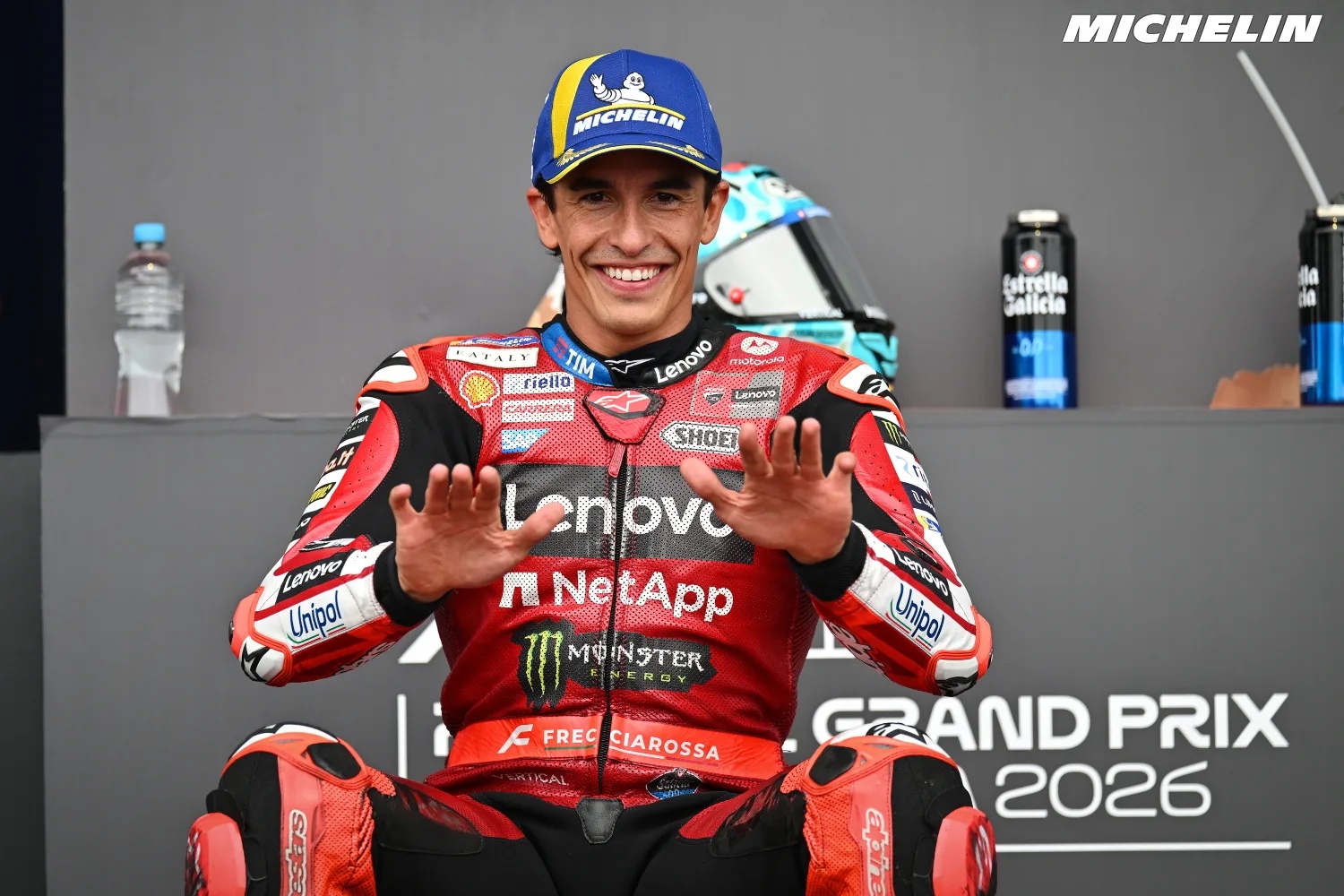 A MotoGP rider in red Ducati leathers smiling and gesturing with both hands at a press event, Grand Prix 2026 backdrop