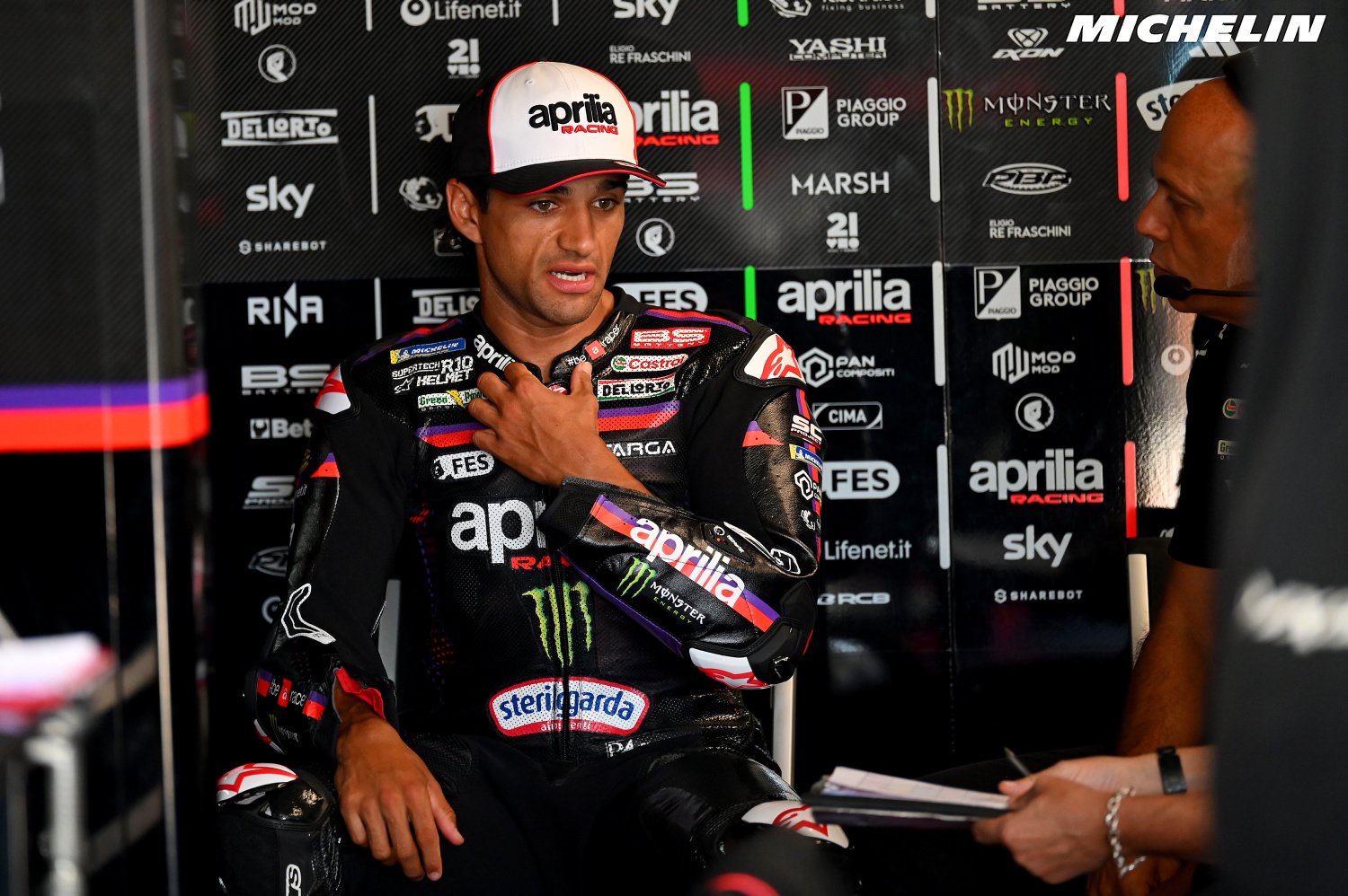 Aprilia Racing rider in black and red leathers with Sterilgarda and Monster logos, seated in the team garage, gesturing with his hand to his chest.