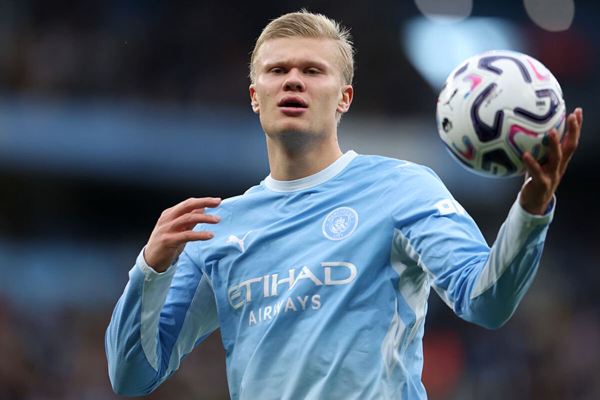 Erling Haaland in Manchester City light blue jersey holding a football, looking forward on the field