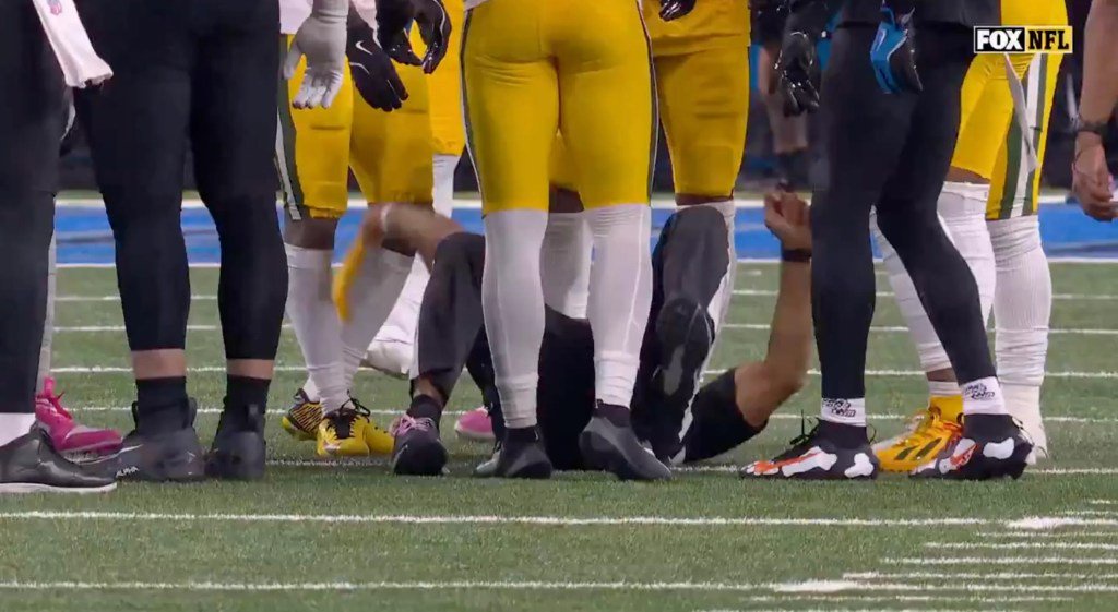 A football player is lying on his back on the field, surrounded by standing players.