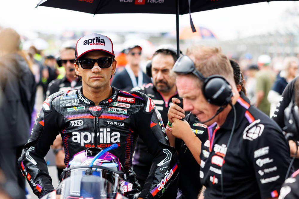 A rider in Aprilia Racing leathers and a red-and-white cap stands in the MotoGP paddock beside a black umbrella, with a helmet and crew members nearby.