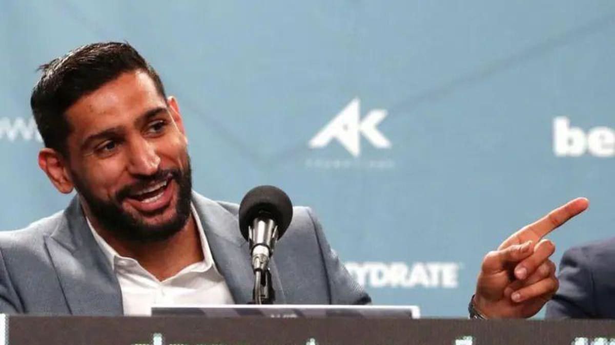Khan smiles and gestures with his lefthand while speaking into a microphone at a press conference. He has short dark hair and a short trimmed beard.