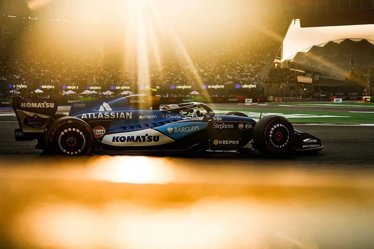 Williams Formula 1 car in Komatsu-sponsored blue and white livery racing at sunset with sun flares, Atlassian and Barclays logos visible