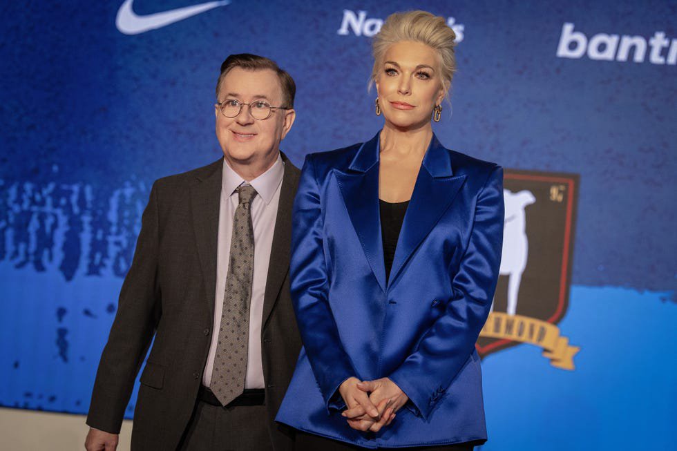 Two individuals pose together at an event with a blue backdrop featuring logos and text.