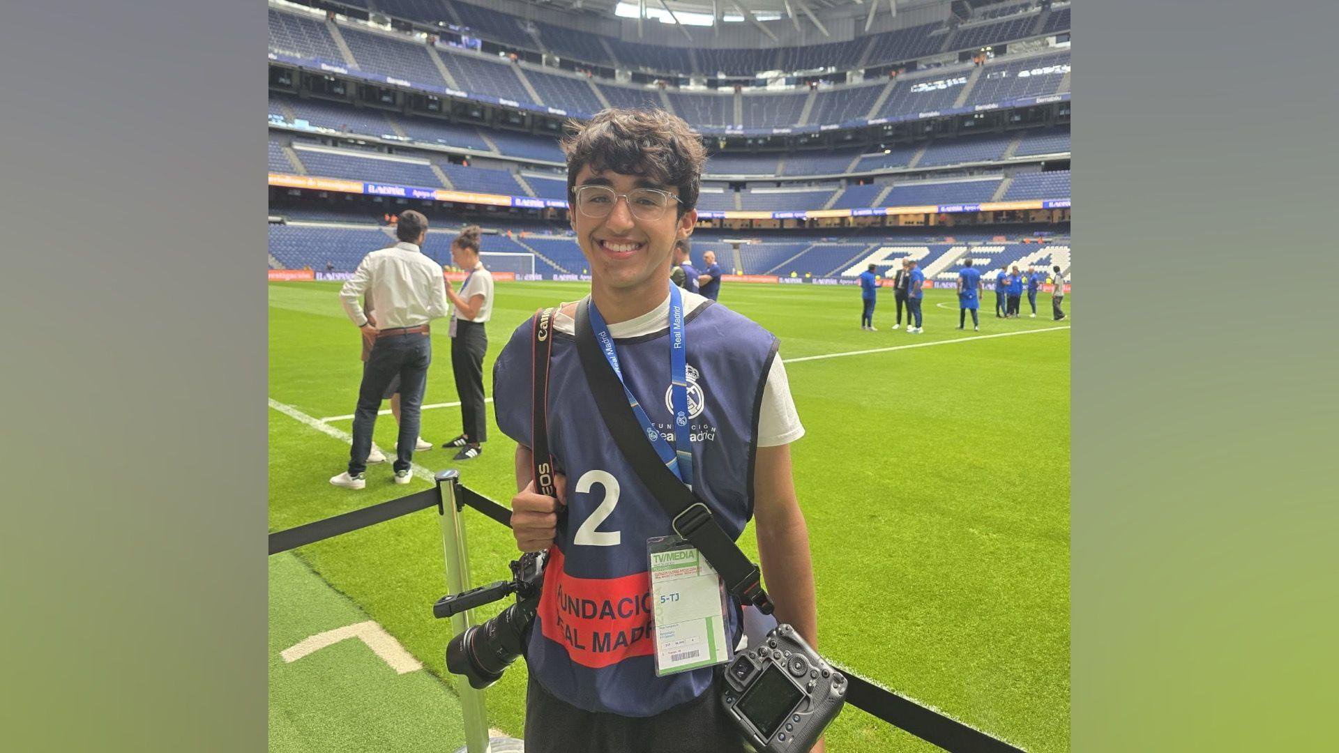 Tiego Greno stands next to the pitch at Real Madrid's Bernabeu stadium. He is wearing a white T-shirt with a blue vest on top which identifies him as a photographer. He has two cameras around his neck. The pitch is behind him, with the five-tiered seating beyond it.
