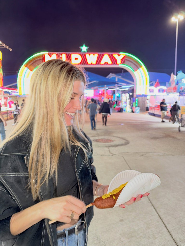 Eugenie Bouchard smiling while holding a corndog at night at a carnival with a neon "MIDWAY" sign in the background.