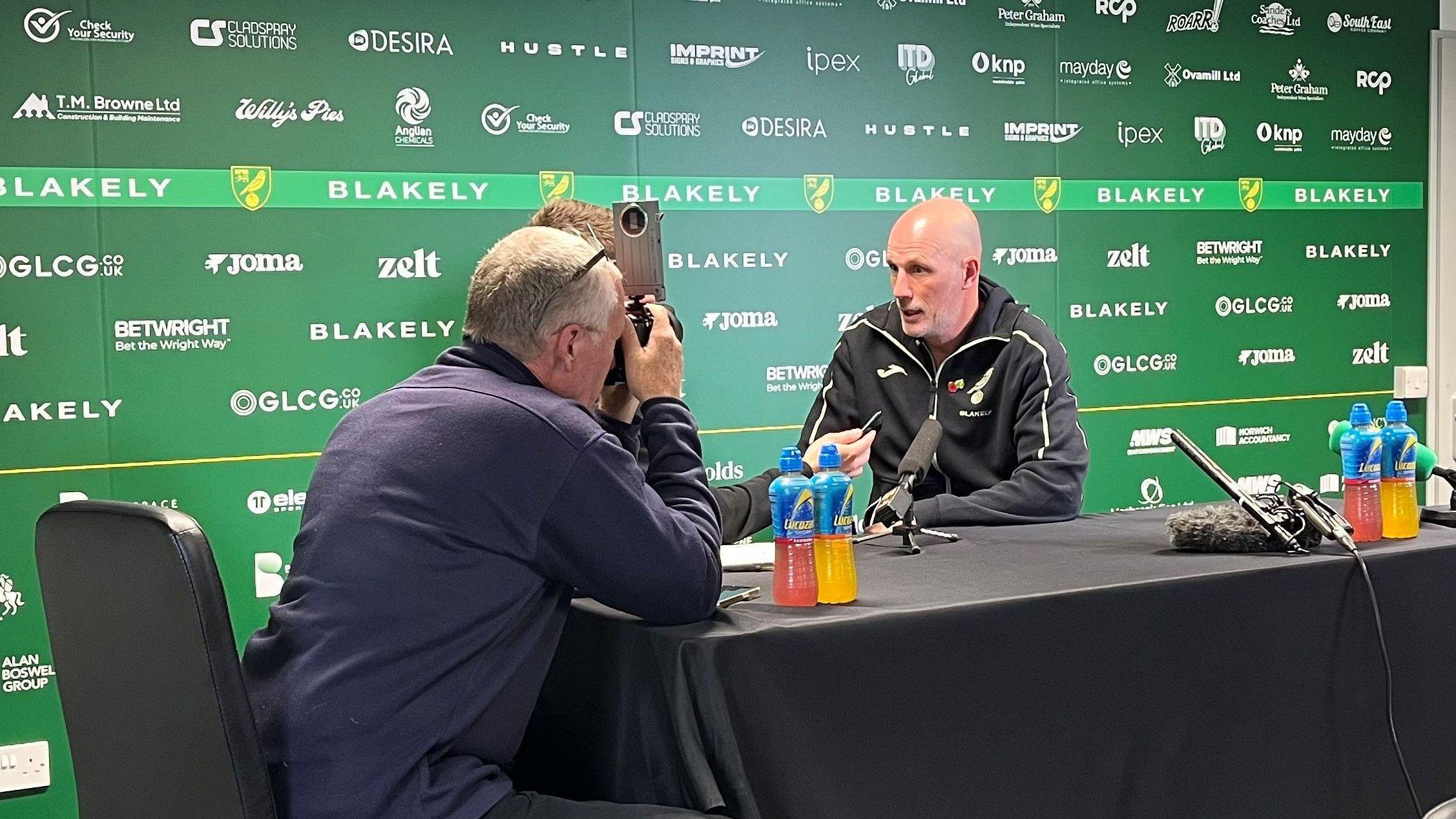 Philippe Clement talks to the media in a press conference at Colney. He's wearing a black training kit leaning on a table with a black table cloth with sports energy drinks dotted around. There's a green hoarding behind with sponsors' logos. A cameraman wearing a blue jumper is filming him.