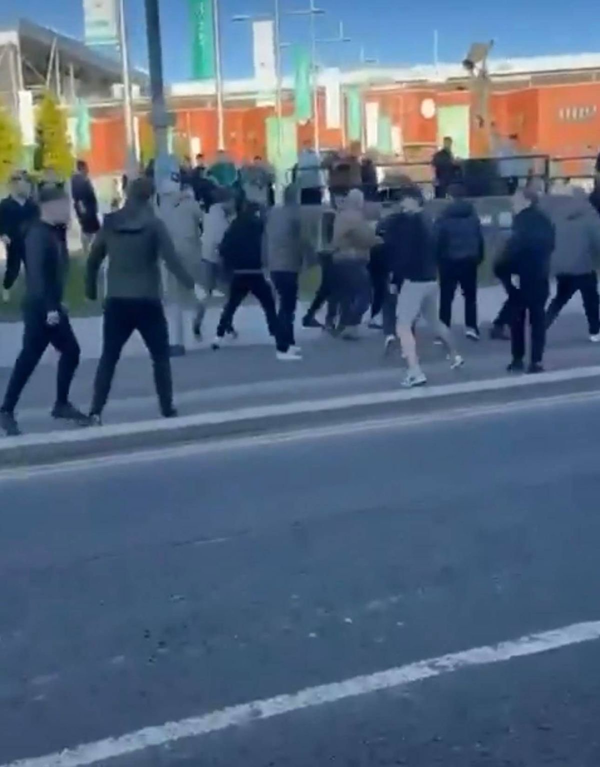 A crowd of individuals are walking swiftly along a street, with a prominent building featuring orange facades in the background.