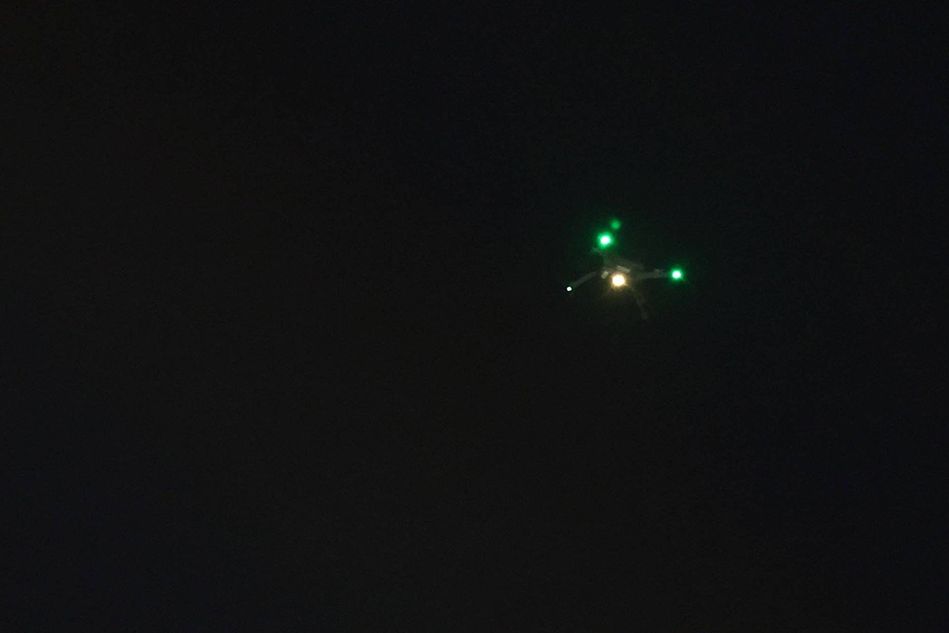 Eine Drohne mit grünen Lichtern fliegt nachts über einem dunklen Himmel.