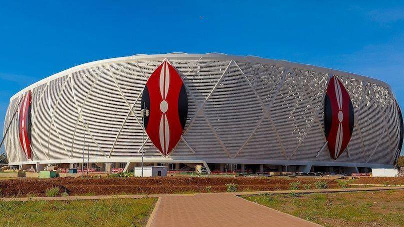 A wide shot of the outside of a football stadium showing three giant Kenyan shields in red, white and black on top of a white wraparound exterior. A clear blue sky is visible above the stadium while partially finished groundsm with scrubby grass, are being worked on in the foreground