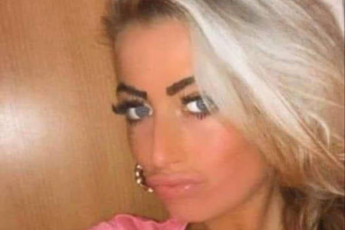 An individual with blonde hair and striking blue eyes, adorned with makeup, is captured in a close-up shot, showcasing a pink top and a wooden background.
