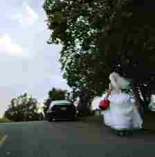 A person in a wedding dress holding a red bag is running down a road with trees and a parked black car in the background