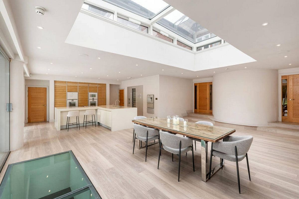 The image showcases an expansive, well-lit room featuring a high ceiling with skylights, polished wooden flooring, and a large dining table surrounded by chairs. The room is predominantly empty, with minimalistic decor and ample natural light.
