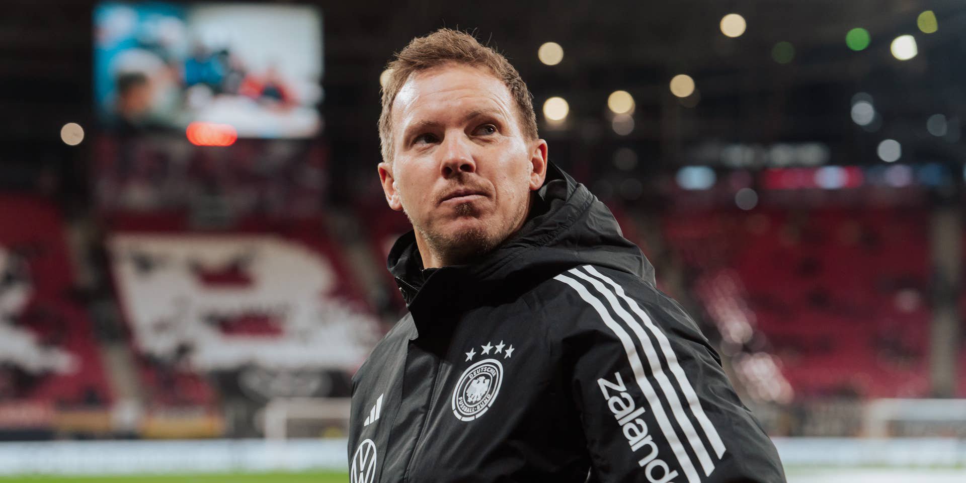Ein Fußballtrainer im Deutschland-Trainingsanzug blickt konzentriert im Stadion nach oben.