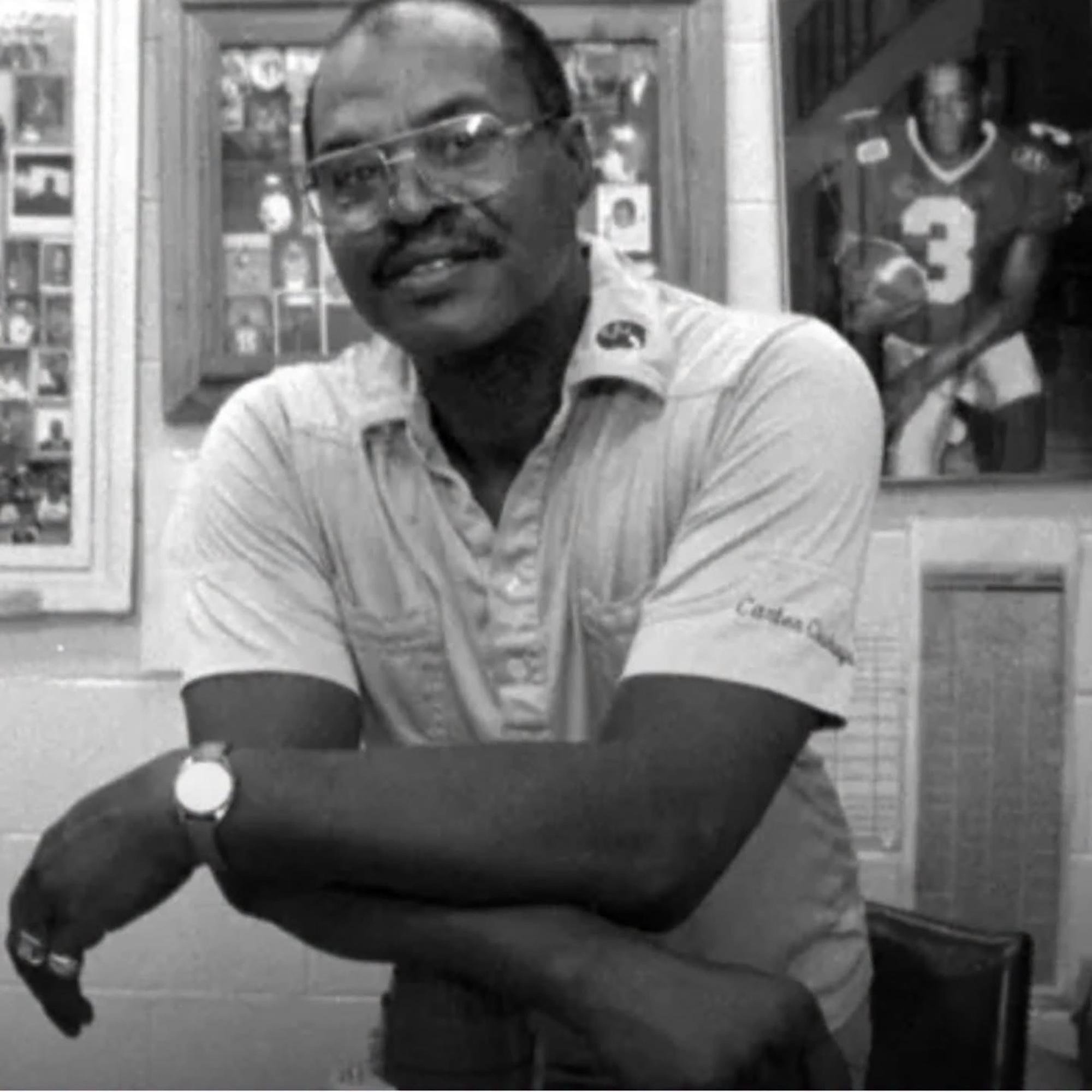 Coach Freddie James leaning on a desk with framed sports photos in the background.