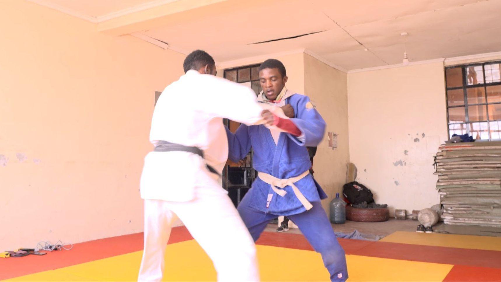 Arno Yafunga Botay grapples with an opponent on a yellow and red judo mat in a sparsely-furnished room. Arno, who is facing the camera, wears a blue judo uniform with a white belt, while his opponent, who has his back to the lens, has a white uniform and a black belt. A pile of smaller mats can be seen on the far right of the image, stacked below a window
