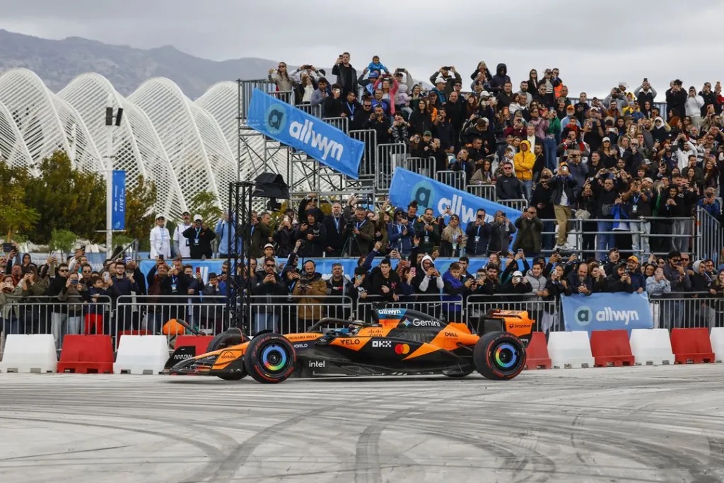 A Formula 1 racing car in orange and black livery performs a demonstration in front of a large, cheering crowd in Athens.