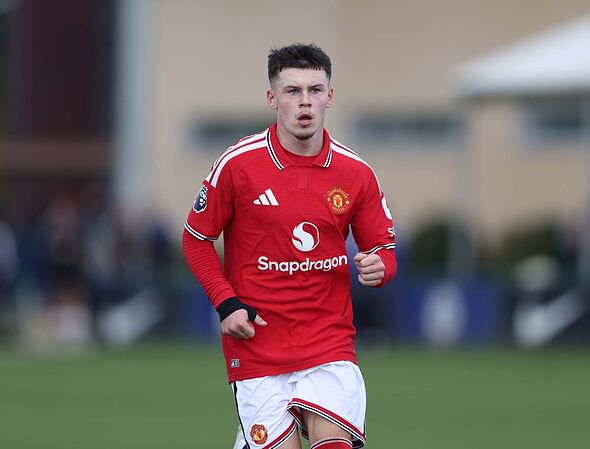 An individual wearing a red football jersey with white stripes and the Adidas logo is running on the field, with a building in t