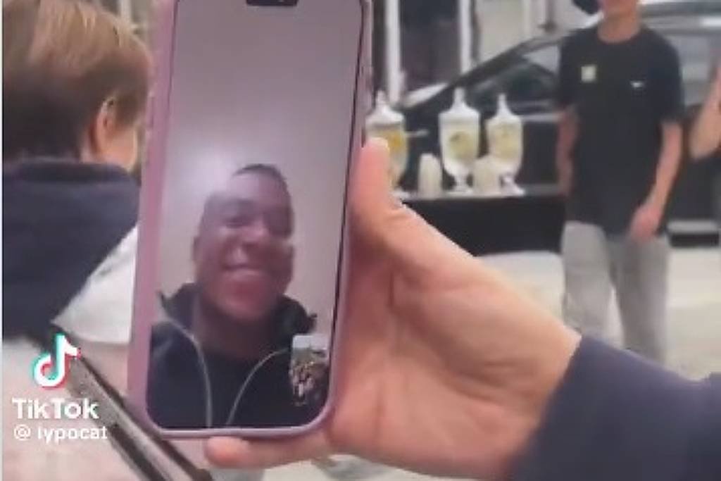 Mão segura celular com chamada de vídeo mostrando rosto sorridente de homem. Ao fundo, pessoas e carros em área urbana com prédios de vidro.