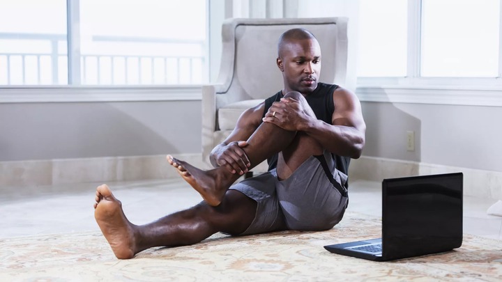 A mid adult African-American man in his 30s exercising at home, taking an online exercise class. He is sitting on the floor doing stretching exercises, while he looks at his laptop. He is an athlete with a muscular build.