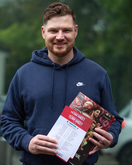 Chris Newman holds souvenir programmes. He is 33 with short reddish hair and beard, and wears a dark blue hoodie. 