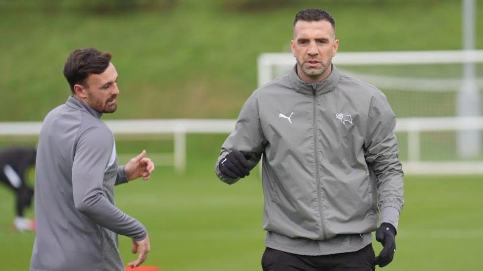 Two footballers wearing grey tracksuits and gloves at a training ground.