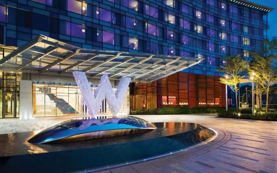 Hotel moderno com fachada de vidro azul, cobertura de vidro na entrada e fonte em forma de W.