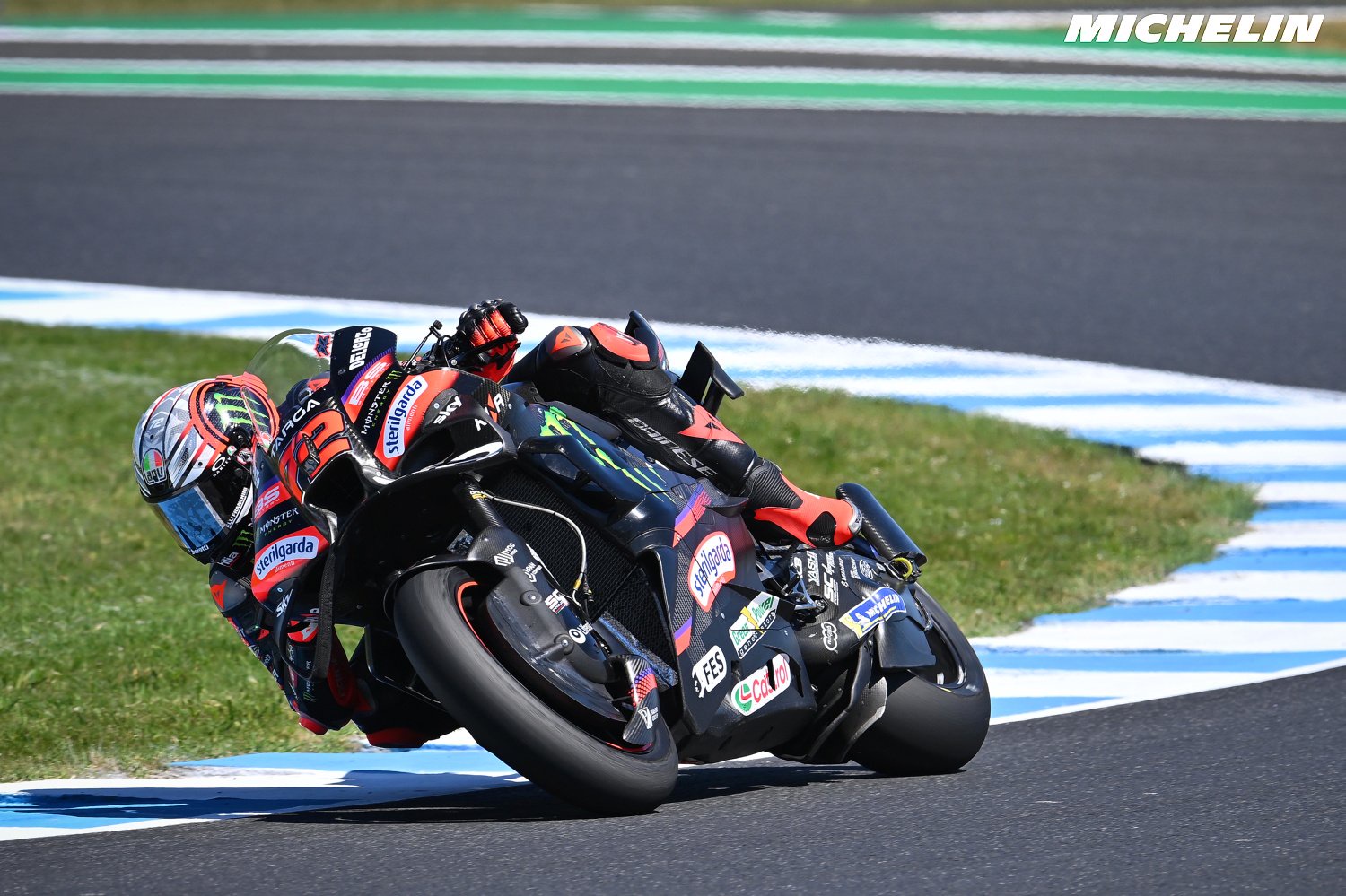 Rider in black and red Aprilia leathers and helmet leaning deep into a corner on a black Aprilia MotoGP bike at Phillip Island, Michelin logo visible