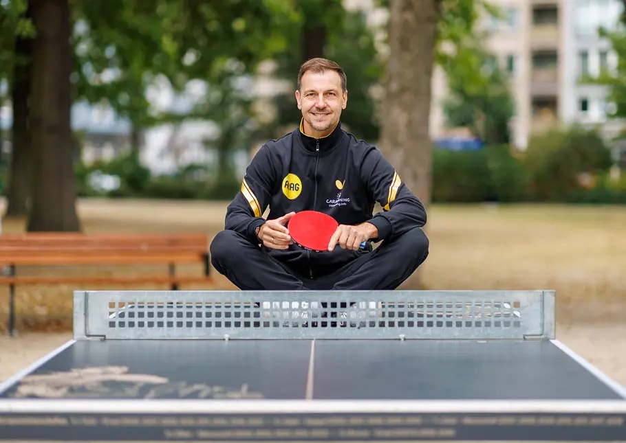 Timo Boll im Trainingsanzug sitzt im Schneidersitz auf einer Tischtennisplatte im Park und hält einen roten Schläger.