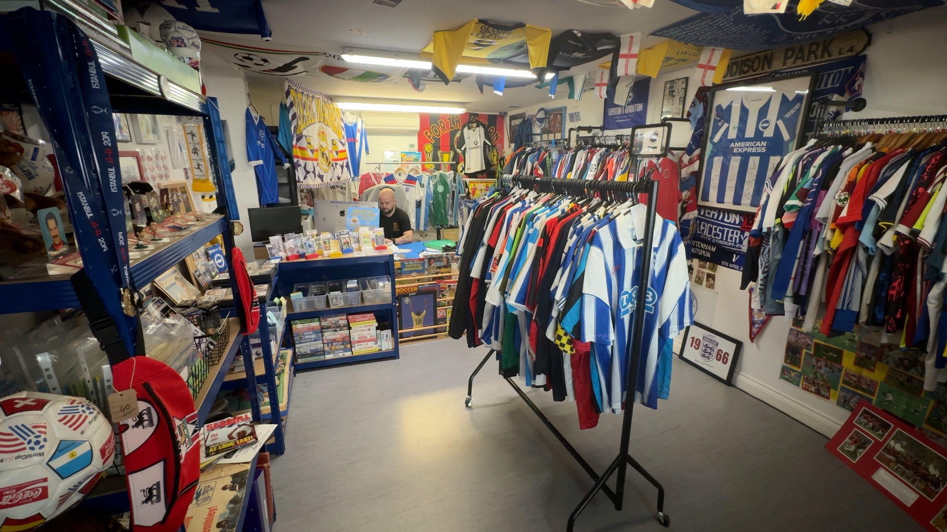 A bald man wearing a black t-shirt is sitting behind the till in a shop filled with football collectibles including a rail full of blue and white Brighton shirts in the centre, shelves full of programmes and football cards on the left, and more rails full of vintage shirts from other clubs on the right wall