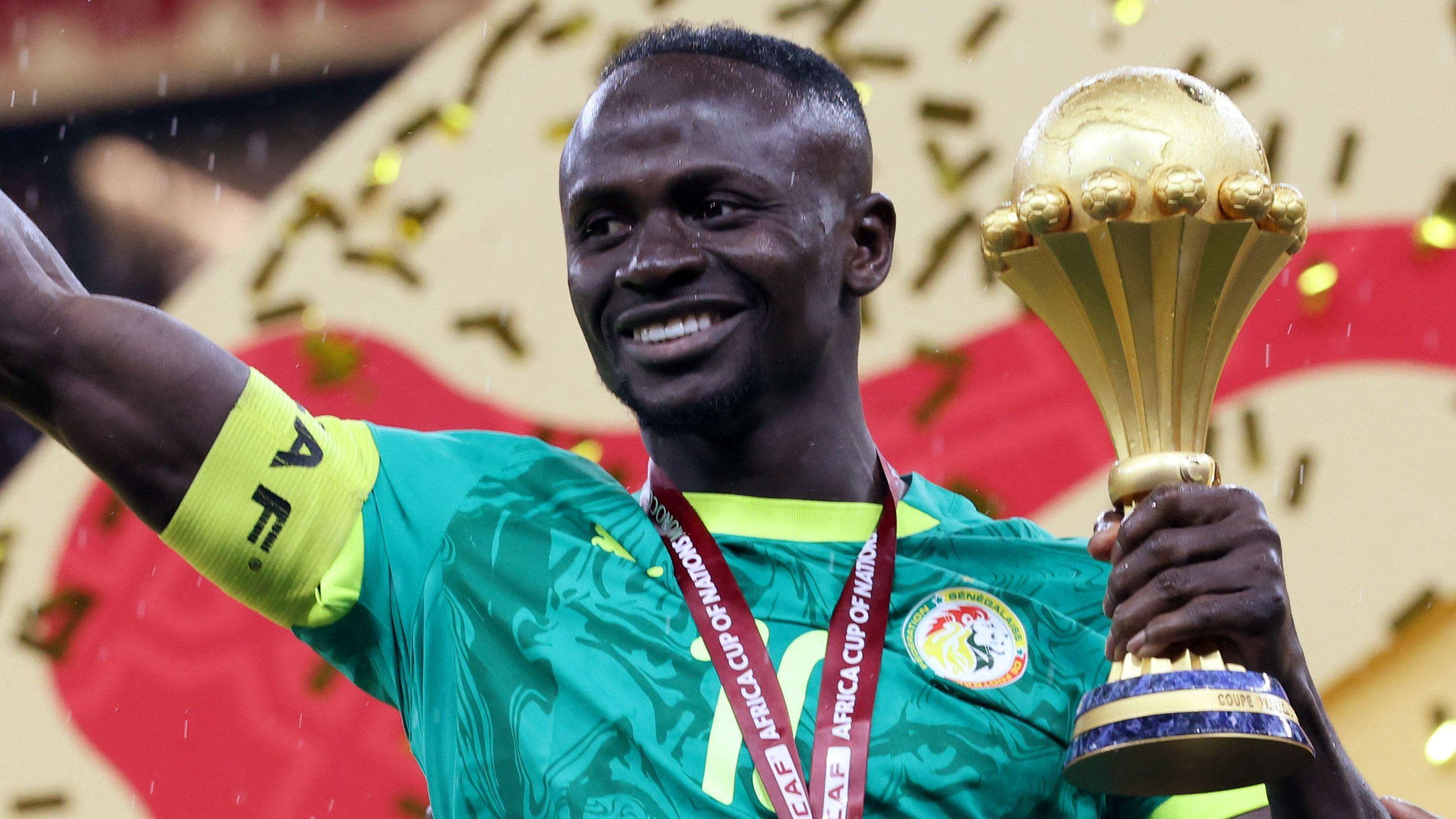 Sadio Mane, visible from chest up wearing a green Senegal shirt and with a yellow captain's armband on his right bicep, smiles as he holds the Africa Cup of Nations trophy in his left hand after the trophy presentation at Afcon 2025 in Morocco. He has a red ribbon around his neck, although the medal is out of shot, and golden ticker tape can be seen falling behind him