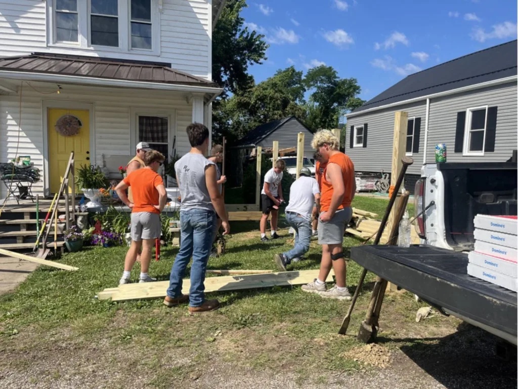 Volunteers set wooden posts in the front yard of a white house as the ramp frame takes shape