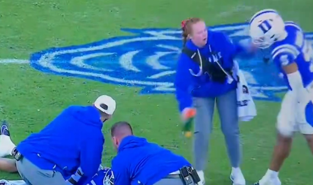 A female athletic trainer is accidentally struck in the face by a football player's arm on the field while medical staff attend to another player.