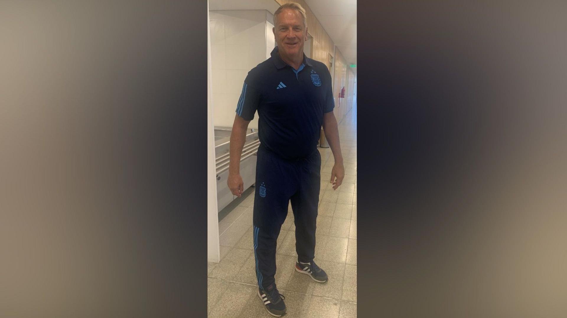 Colin is wearing the Argentinian kit and standing inside a sporting facility smiling at the camera. The kit is navy blue wit lighter blue detail.