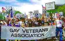 A group of men and women hold signs and wave flags as they march down a street in Washington, D.C. A few people at the front of the crowd, leading the rest, hold up a large banner that reads "Gay Veterans Association."
