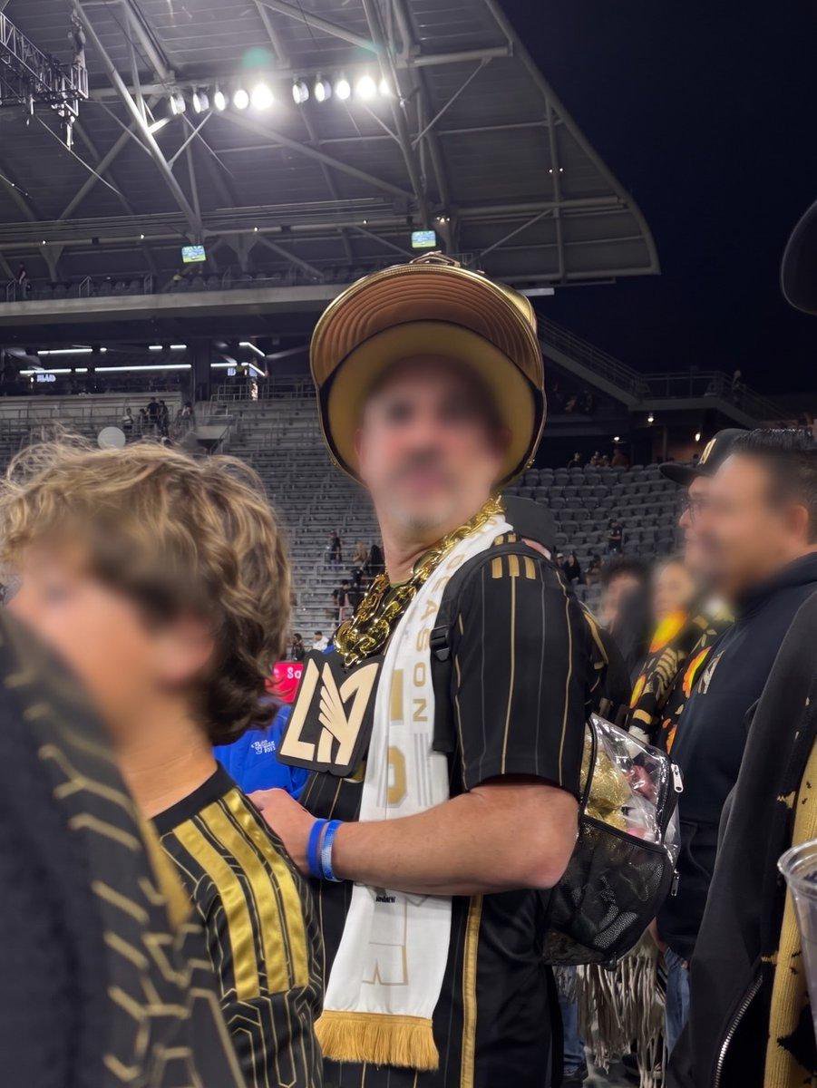 A fan wearing an oversized gold hat and an LAFC jersey stands in the stands of a stadium.