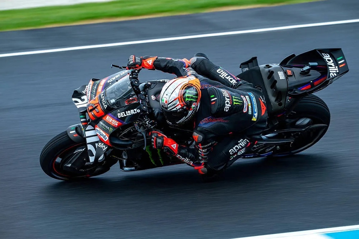 Rider in black Aprilia leathers and helmet leaning deep into a corner on a wet MotoGP track, with Aprilia and Sky branding visible.
